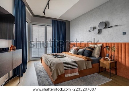 Interior design of elegant bedroom with big orange bed, beige and grey bedclothes, blue curtain, rug, modern lamp, night stand, vase with dried flowers and personal accessories. Home decor. Template. Royalty-Free Stock Photo #2235207887