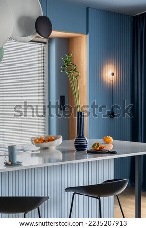 Interior design of kitchen interior with marble kitchen island, blue wall, black chokers, bowl with fruits, big window, cup, wooden floor, lamp on wall and personal accessories. Home decor. Template. Royalty-Free Stock Photo #2235207913