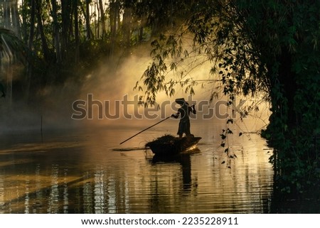 Beautiful view of an asian adult male old boatman rowing a wooden boat with a bamboo stick across a small river stream during sunset to deliver dry grasses for animal feeds in northeast Thailand Royalty-Free Stock Photo #2235228911