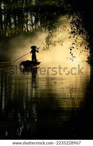 Beautiful view of an asian adult male old boatman rowing a wooden boat with a bamboo stick across a small river stream during sunset to deliver dry grasses for animal feeds in northeast Thailand Royalty-Free Stock Photo #2235228967