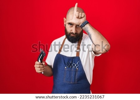 Young hispanic man with beard and tattoos wearing barber apron holding razor making fun of people with fingers on forehead doing loser gesture mocking and insulting. 