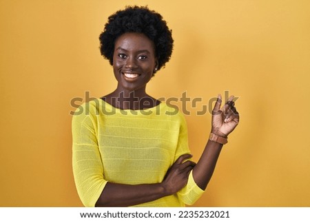 African young woman standing over yellow studio with a big smile on face, pointing with hand finger to the side looking at the camera. 