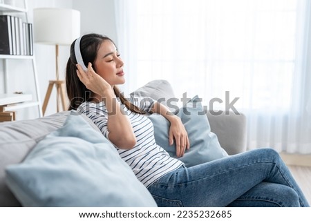 Asian woman listen to music and dancing while sitting on sofa house. Attractive beautiful young girl put on headphone feeling happy to spend leisure time at home and enjoy weekend activity at home. Royalty-Free Stock Photo #2235232685