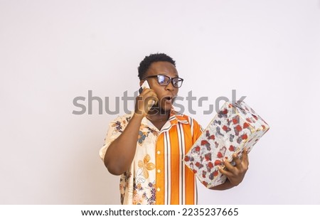 surprised African American male with glasses on a white background holding a gift box while making a phone call