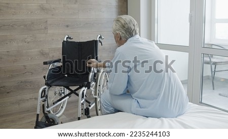 Unhappy elderly man looking at wheelchair in a hospital room, disability, reduced mobility