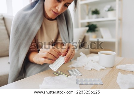 A woman with a cold with pills is treated at home chooses which drugs to take and self-medicates, checks the expiration date while sitting on the couch at home Royalty-Free Stock Photo #2235256125
