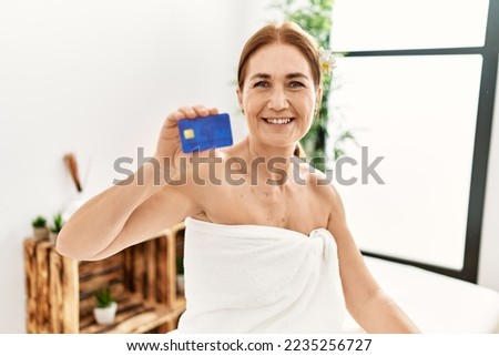 Middle age caucasian woman wearing towel holding credit card sitting on massage board at beauty center