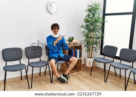 Young hispanic man sitting at doctor waiting room with neck injury amazed and smiling to the camera while presenting with hand and pointing with finger. 