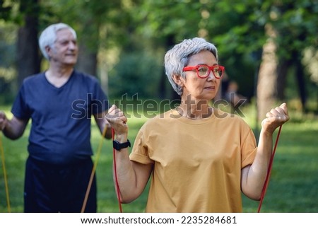 Senior 60 spouses wear sportswear doing hands exercises, training arms outdoor in summer park in the morning using resistance rubber bands. Healthy lifestyle, active retired life use modern appliance Royalty-Free Stock Photo #2235284681