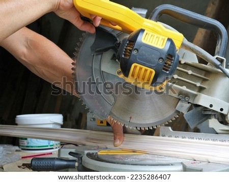 working on a circular saw in a workshop, male muscular hands lower the handle and blade of a miter saw to cut off the edge of a wooden fillet Royalty-Free Stock Photo #2235286407