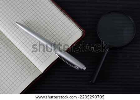 An open checkered notebook, a silver fountain pen and a black magnifying glass lie on a dark textured surface. Concept of research, investigation and information analysis. copyspace. Close-up