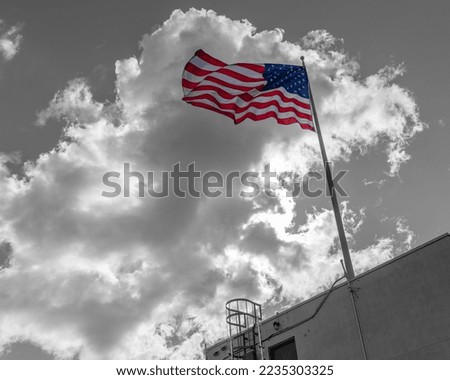 A large colorful American flag blows in the wind against a cloudy monochrome
 sky.