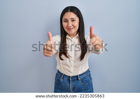 Young latin woman standing over blue background approving doing positive gesture with hand, thumbs up smiling and happy for success. winner gesture. 