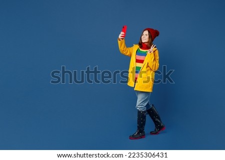 Full body young woman wear sweater red hat yellow waterproof raincoat doing selfie shot on mobile cell phone isolated on plain dark royal navy blue background Outdoors wet fall weather season concept