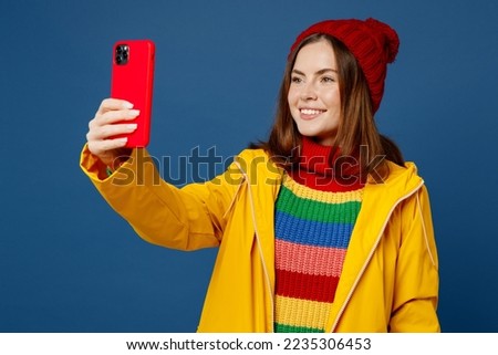 Young woman wear sweater red hat yellow waterproof raincoat outerwear do selfie shot on mobile cell phone post isolated on plain dark royal navy blue background Outdoor wet fall weather season concept