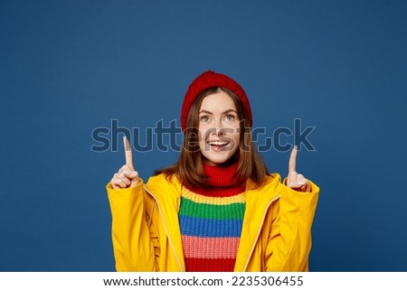 Young fun woman wear sweater red hat yellow waterproof raincoat outerwear point index finger overhead on area isolated on plain dark royal navy blue background Outdoors wet fall weather season concept