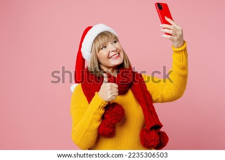 Merry elderly woman 50s years old wear yellow knitted sweater red scarf Santa hat posing doing selfie shot on mobile cell phone isolated on plain pink background. Happy New Year Christmas 2023 concept