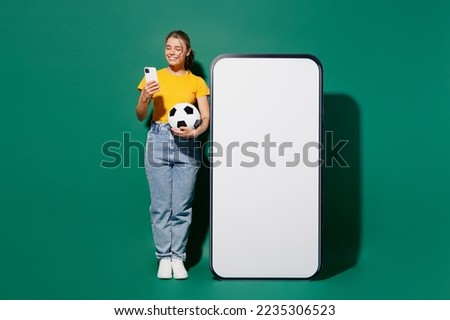 Full body young woman fan wear yellow t-shirt cheer up support football sport team hold soccer ball watch tv live stream big blank screen mobile cell phone use smartphone isolated on green background