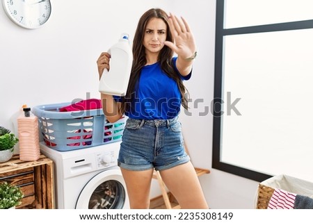 Young brunette woman doing laundry holding detergent bottle with open hand doing stop sign with serious and confident expression, defense gesture 