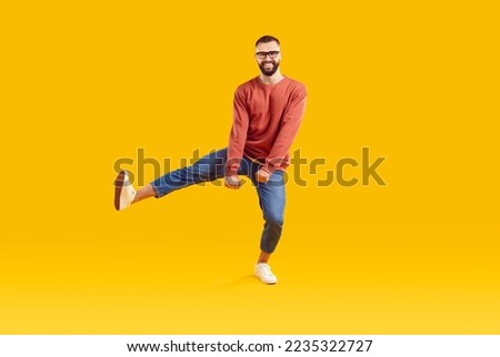 Smiling funky cheerful young bearded man with short hair in glasses wearing red sweatshirt and denim pants dancing on one leg isolated on yellow background. Good mood happy lifestyle nice day concept. Royalty-Free Stock Photo #2235322727