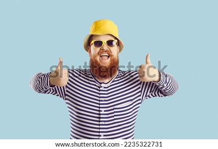 Portrait of cheerful funny chubby man showing thumbs up isolated on pastel light blue background. Red-bearded man in panama, sunglasses and striped clothes with funny expression recommends something.