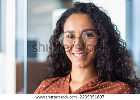 Close up photo portrait of beautiful Latin American woman with curly hair and glasses, businesswoman inside office building smiling and looking at camera.