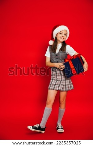 full length of cheerful child in plaid skirt and santa hat holding blue gift box on red background