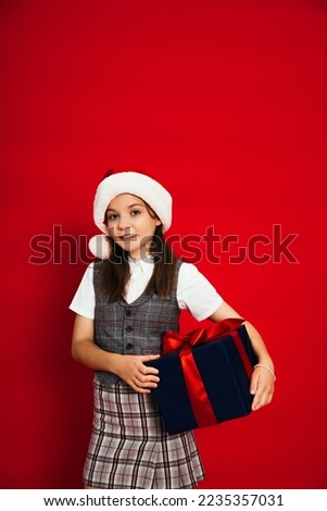 pleased girl in checkered vest and santa hat holding gift box isolated on red