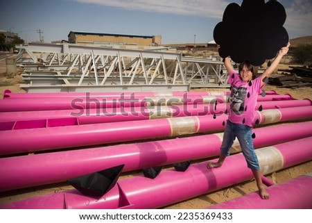 a cute kid in a pink t-shirt with a cat print plays among pink pipes with black clouds, rain, boats and paper planes against the blue sky in an industrial location. fashion style photoshoot