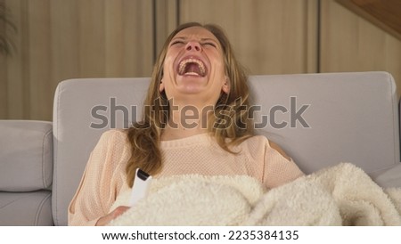 CLOSE UP: Beautiful woman enjoying on a comfy sofa and laughing at funny comedy. Pretty lady covered with blanket and eating snacks while watching movie. Young woman at relaxing on a winter evening.