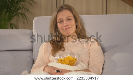 CLOSE UP: Beautiful woman getting emotional at watching romantic movie on TV. Pretty lady covered with blanket and eating snacks while watching movie. Young woman enjoying on a winter evening.