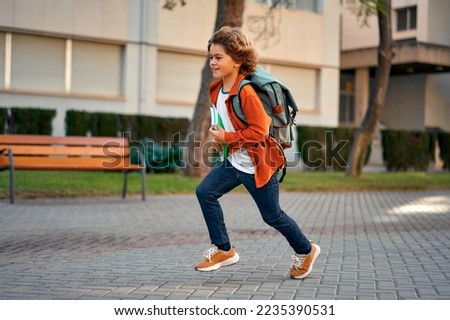 A happy cute smart boy with a stylish hairstyle with a school bag and notebooks in his hand runs late for lessons to school. Modern backpack. First time to school. Back to school. Royalty-Free Stock Photo #2235390531