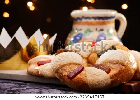 Traditional king cake or rosca with candied fruit by a typical Mexican earthenware cup of hot chocolate and a golden crown over a black background. Celebration of the Three Kings Day or Epiphany. Royalty-Free Stock Photo #2235417157