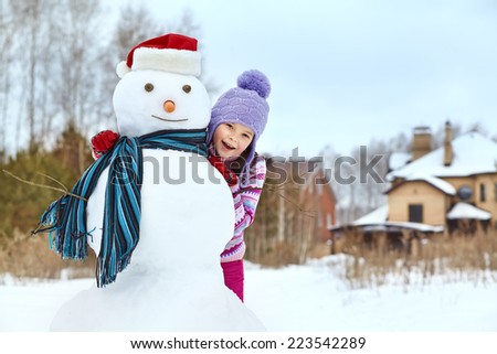 happy kid playing with snowman in santa hat. funny little girl on a walk in the winter outdoors