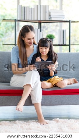 Millennial Asian happy family mother sitting smiling on cozy sofa couch holding showing funny content streaming online via touchscreen smartphone to little young girl daughter in living room at home. Royalty-Free Stock Photo #2235451803