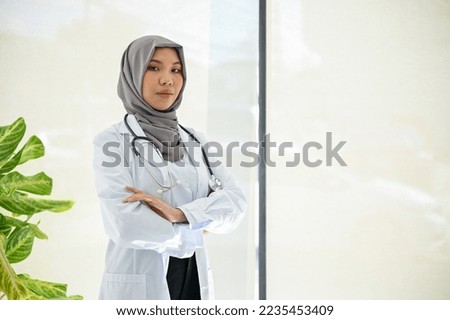 Professional and elegant Muslim female doctor in uniform and hijab stands in the hospital, arms crossed, looking at the camera.