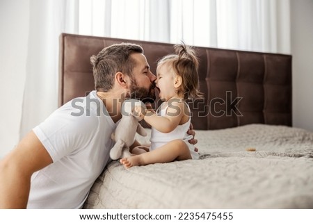 A father is having eskimo kisses with his daughter while she is sitting on a bed. Royalty-Free Stock Photo #2235475455