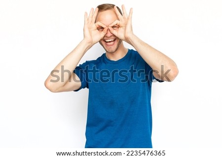Happy Caucasian man showing OK signs over his eyes. Portrait of adult with fair hair smiling at camera, standing on white background, having fun. Happiness, positivity concept