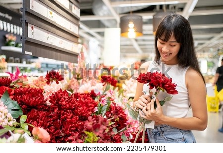 Portrait of an Asian young happy woman in casual choosing fresh seasonal artificial flowers to buy for her house decor. Red rose flowers shop in department store shopping in holiday weekend. Royalty-Free Stock Photo #2235479301