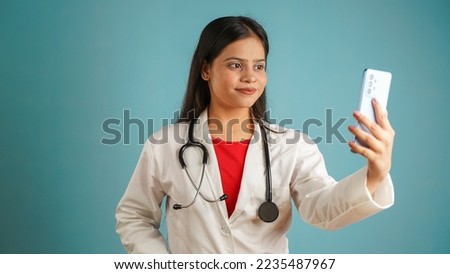 Portrait of a young female doctor taking selfie with mobile phone, Asian Indian woman doctor in apron isolated over blue studio background, taking photo with smartphone