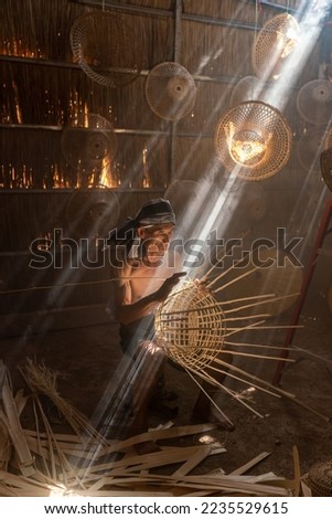 Asian village lifestyle concept. A male asian mature adult worker weaving bamboo basket and woven hat under the beautiful sun rays in a room in Sakon Nakhon, Northeast Thailand, Isan way of life. Royalty-Free Stock Photo #2235529615