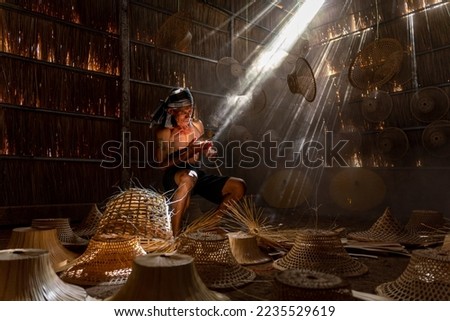 Asian village lifestyle concept. A male asian mature adult worker weaving bamboo basket and woven hat under the beautiful sun rays in a room in Sakon Nakhon, Northeast Thailand, Isan way of life. Royalty-Free Stock Photo #2235529619