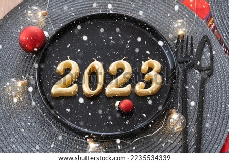Golden figures 2023 from candles on a black plate on a festive table with a New Year's serving. Loft-style interior, party, feast. Wicker napkin, fork, knife