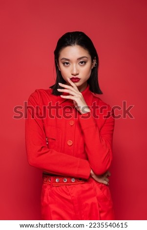 brunette asian woman in fashionable blazer holding hand near face while looking at camera isolated on red Royalty-Free Stock Photo #2235540615