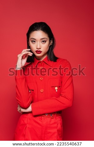 brunette asian woman in fashionable jacket holding hand near face isolated on red