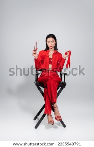 full length of elegant asian woman in red attire holding glove and sunglasses while sitting on grey background Royalty-Free Stock Photo #2235540791