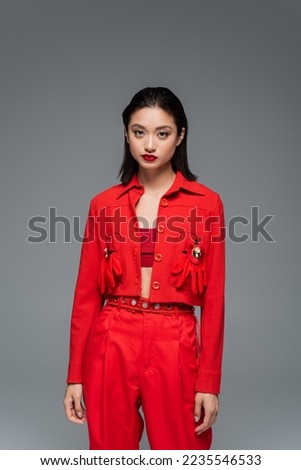 young asian woman in red jacket decorated with brooches and gloves looking at camera isolated on grey Royalty-Free Stock Photo #2235546533