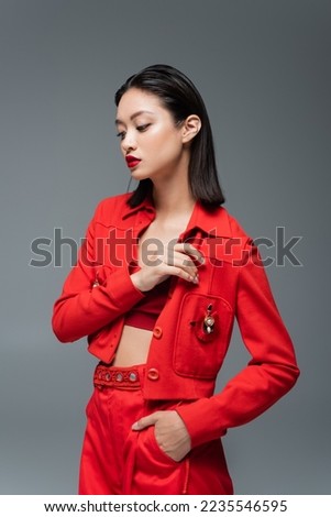 asian woman in red pants and jacket with brooch posing with hand in pocket isolated on grey Royalty-Free Stock Photo #2235546595