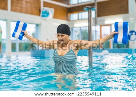 Dedicated elderly caucasian woman in striped swimsuit and black hair cap using pool buoys to do aqua exercised. Swimming pool interior. High quality photo Royalty-Free Stock Photo #2235549111