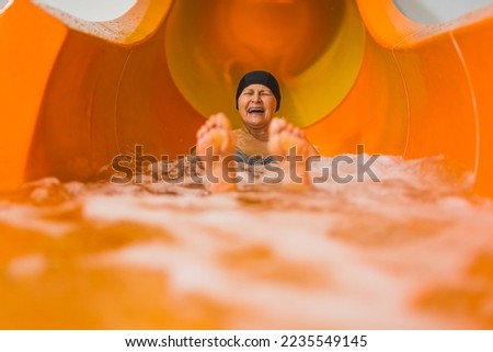 Fun swimming pool or aqua park activities. Laughing amused elderly caucasian female pensioner in black hair cap sliding on an orange water slide. Blurred feet in the foreground. High quality photo Royalty-Free Stock Photo #2235549145